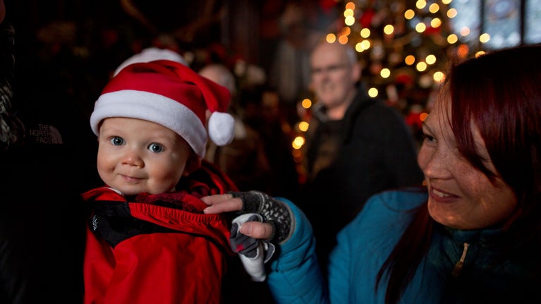 A small child in a Santa hat with their family and Christmas lights in the background
